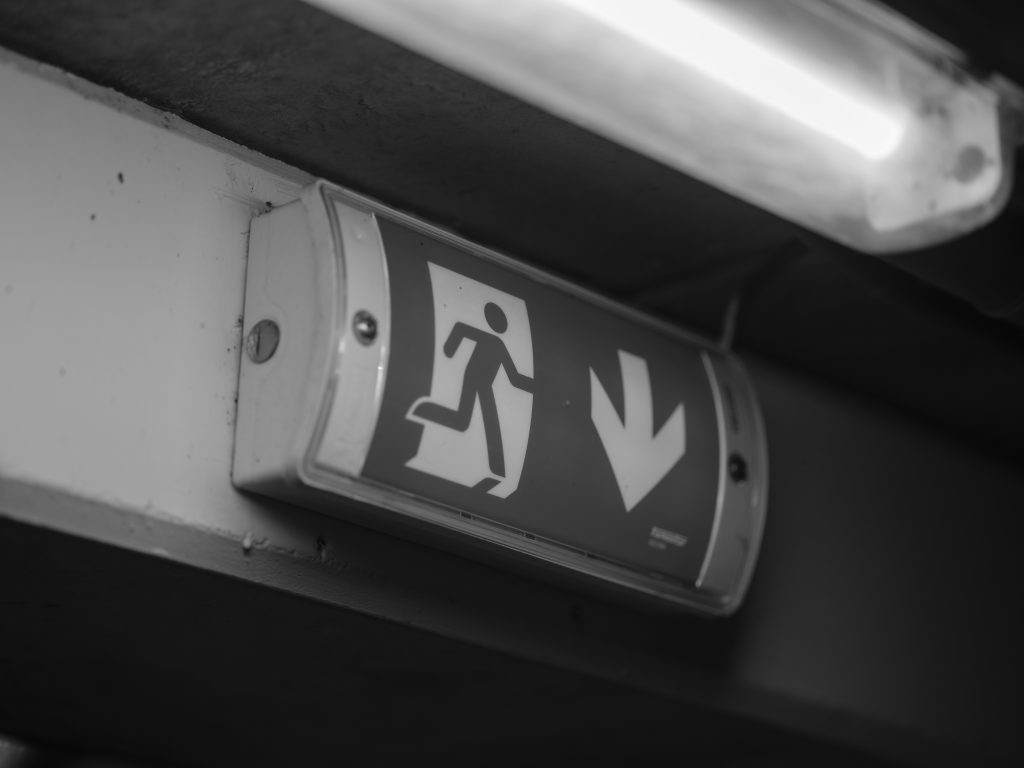 pexels-photo-33863787-33863787 A black and white image of an emergency exit sign in a dimly lit garage with fluorescent lighting.